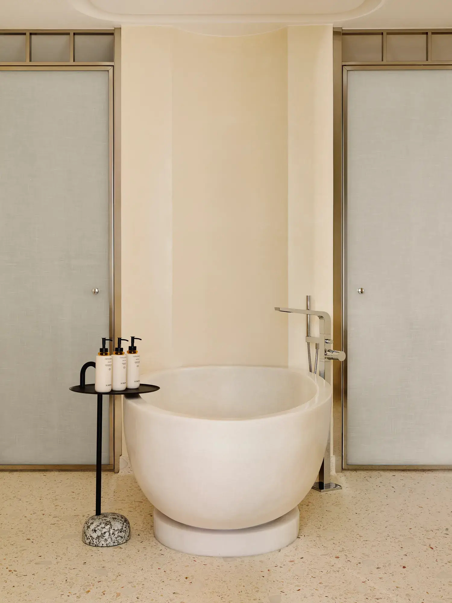 Salle de bain moderne avec baignoire ovale autoportante, robinetterie chromée élégante et petite table avec produits de toilette de luxe.