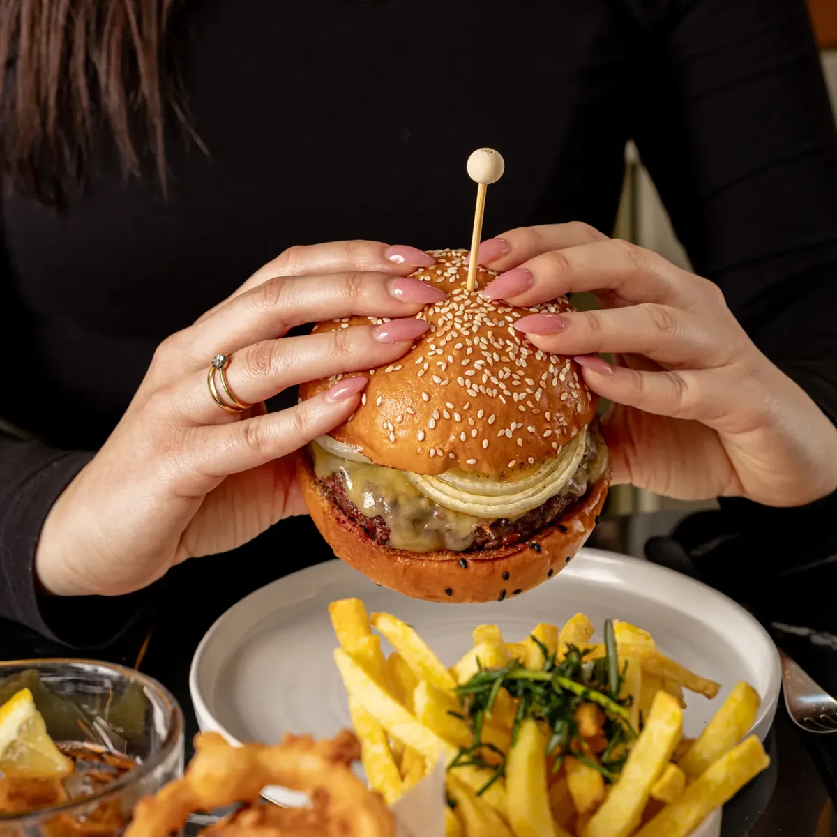 Hands holding a cheeseburger with sesame bun, served with fries and onion rings on a restaurant table.
