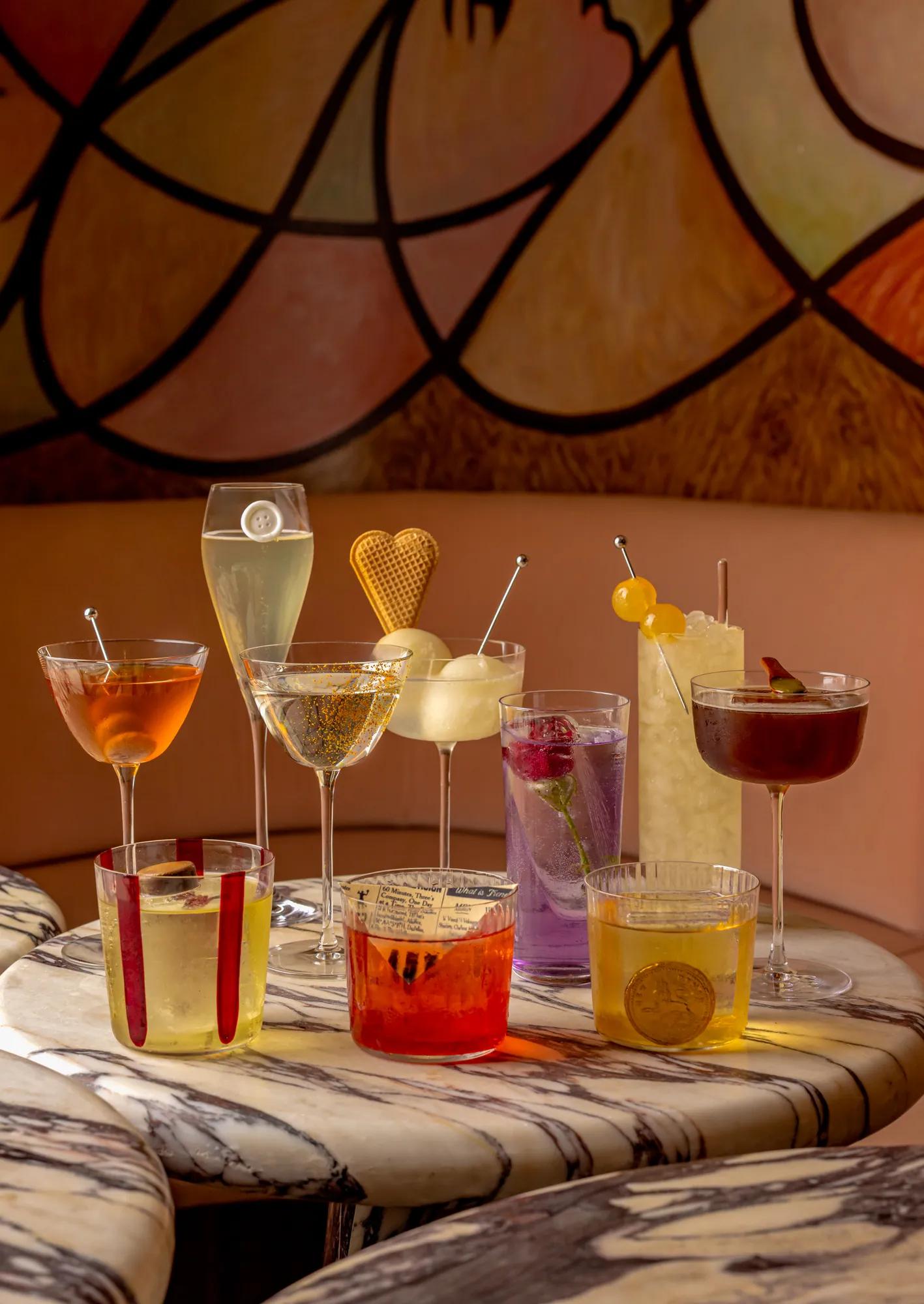 Eight colourful cocktails and champagne glasses arranged on a marble table in a stylish lounge with warm abstract wall art behind.