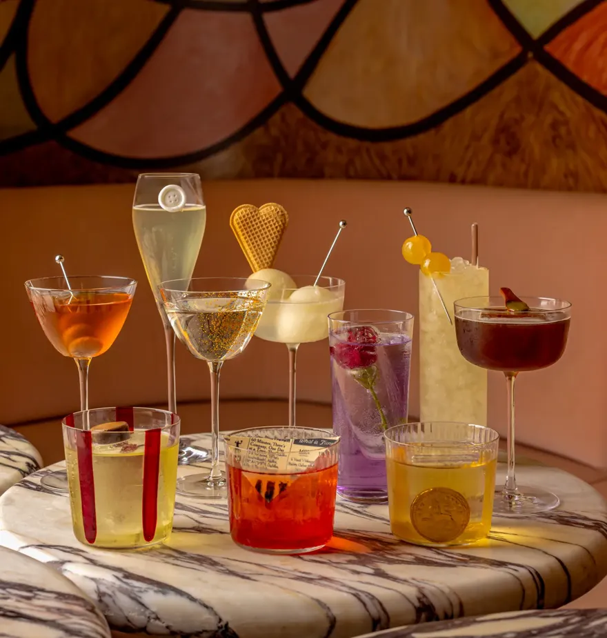 Eight colourful cocktails and champagne glasses arranged on a marble table in a stylish lounge with warm abstract wall art behind.