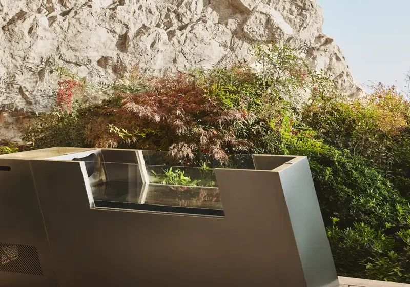 Modern outdoor ice bath beside landscaped greenery and a stone cliff wall.