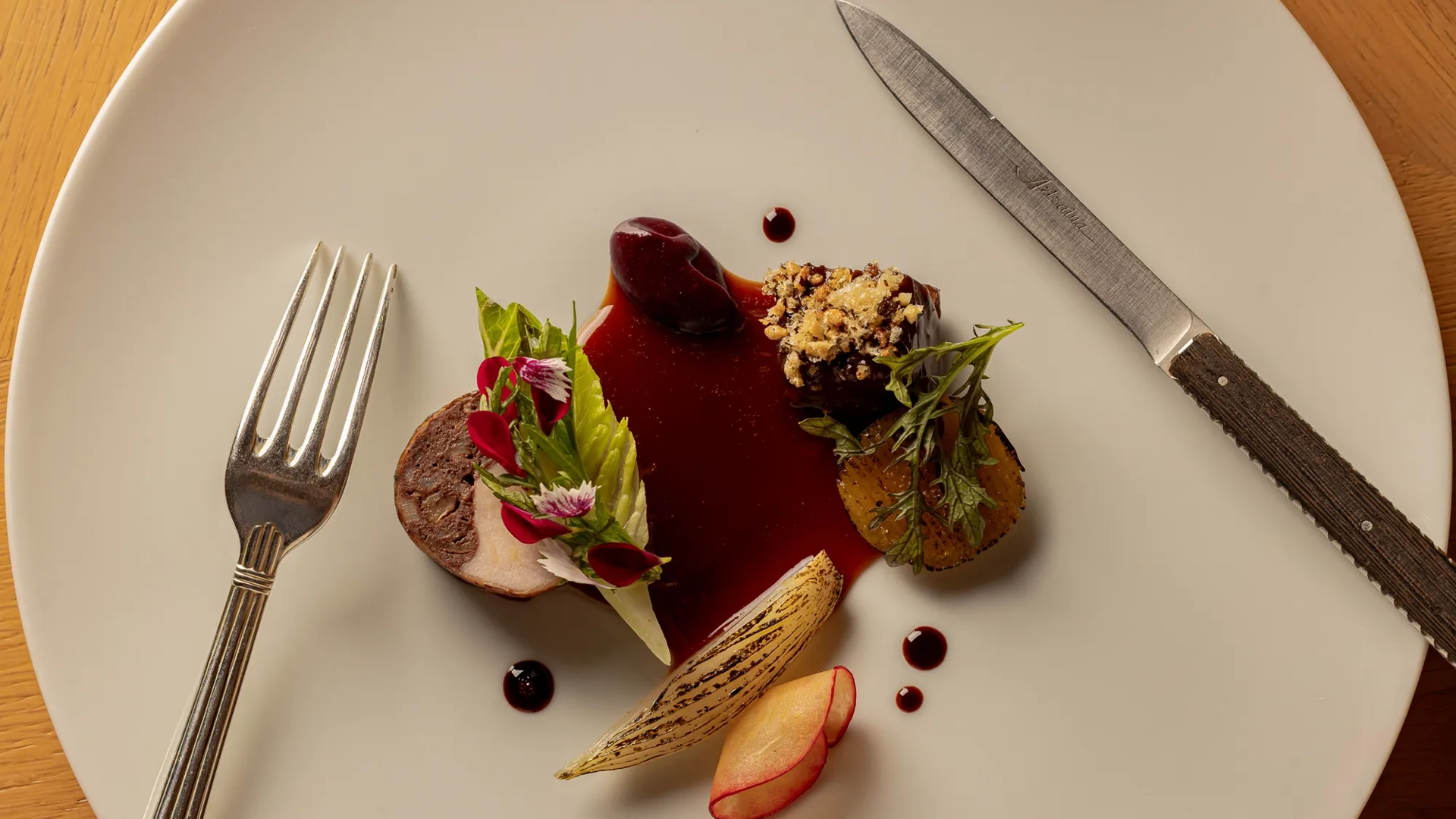 Plated fine-dining dish with sliced meat, vegetables, and a dark sauce, arranged artfully on a large white plate.
