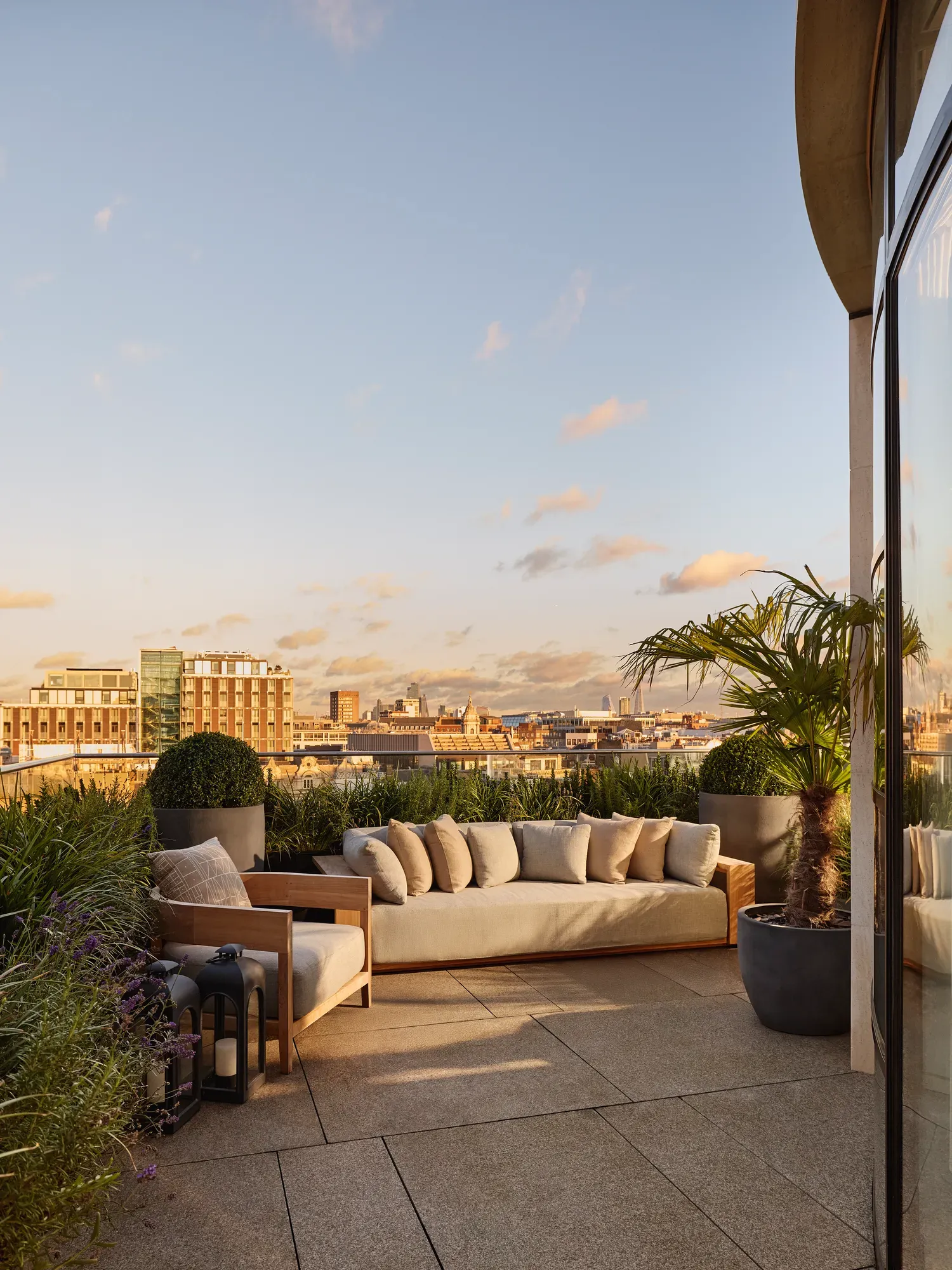 Rooftop terrace at sunset with a cream outdoor sofa, wooden armchair, and potted plants including a palm tree, overlooking the London skyline under a soft golden sky.