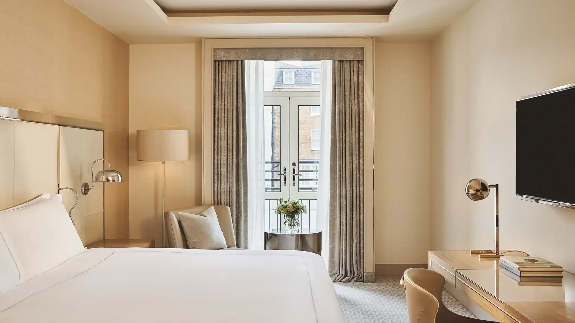 Bright bedroom with king bed, desk and armchair, opening to a small balcony through French doors with city views.