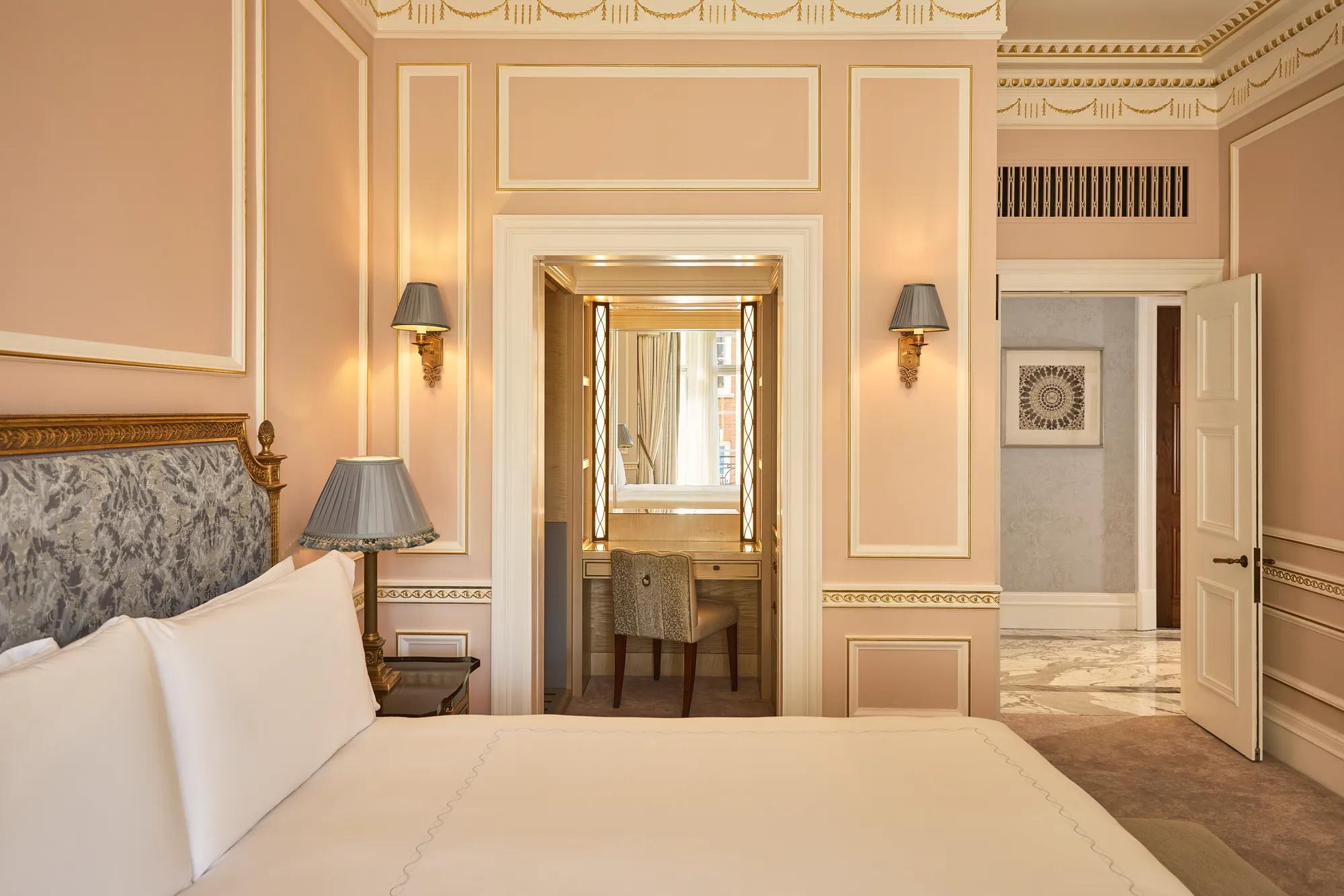 Partially viewed king-size bed in a classically styled bedroom with gold detailing, bedside lamps, and a dressing area leading to a marble-clad entrance hall.