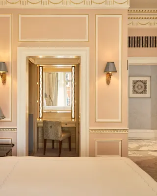 Partially viewed king-size bed in a classically styled bedroom with gold detailing, bedside lamps, and a dressing area leading to a marble-clad entrance hall.