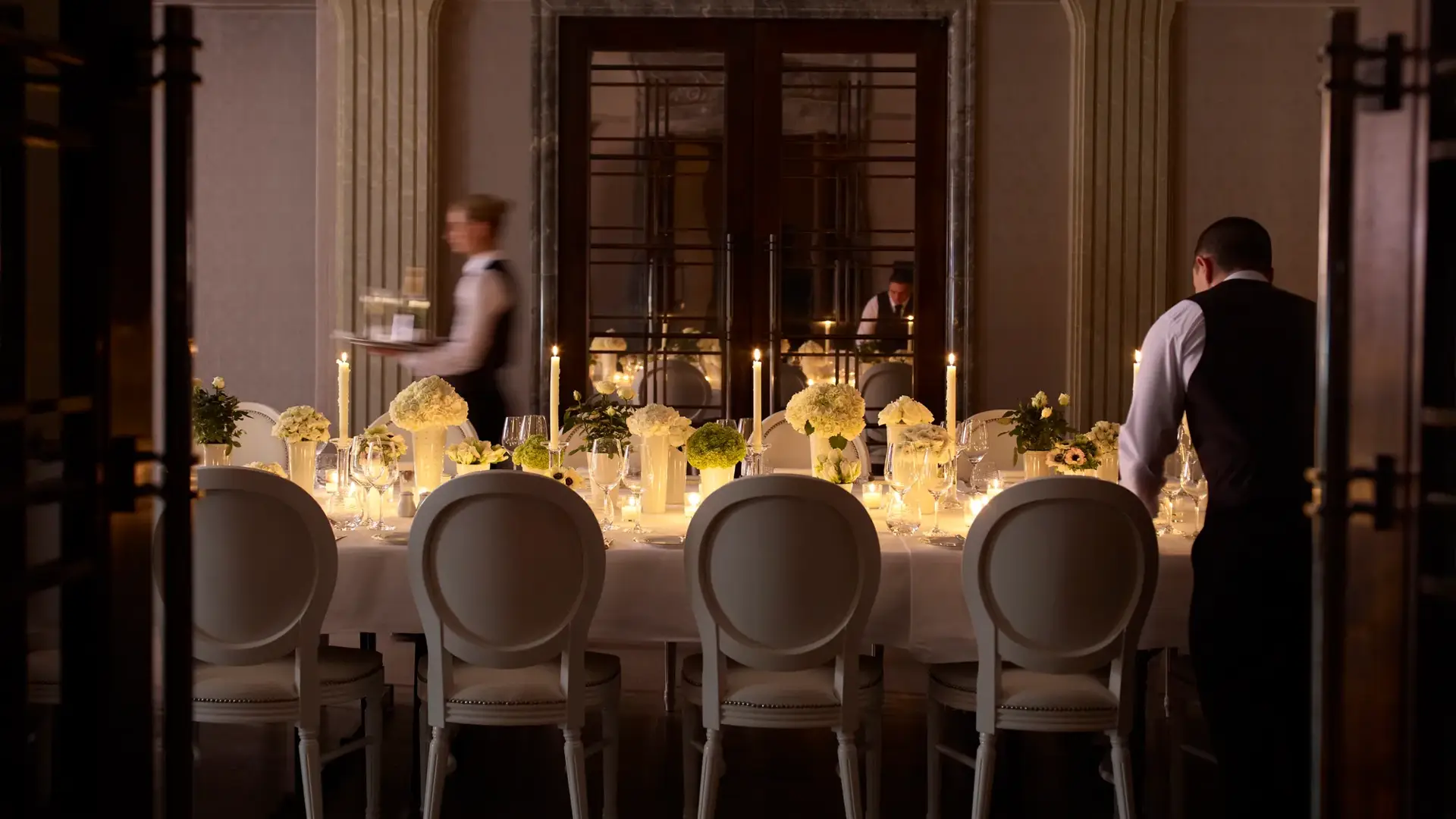Elegant private dining room with a long candlelit table, floral centrepieces, and staff preparing place settings.