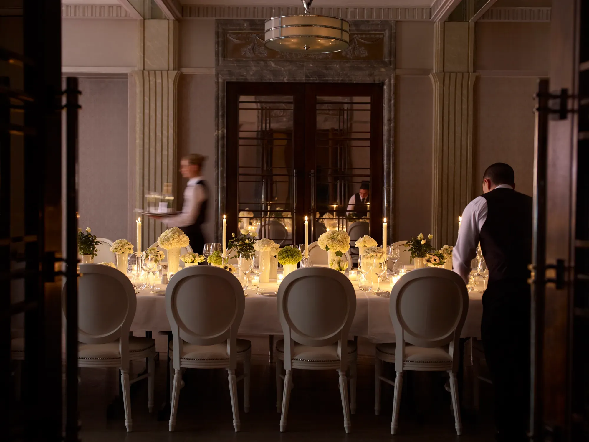 Elegant private dining room with a long candlelit table, floral centrepieces, and staff preparing place settings.