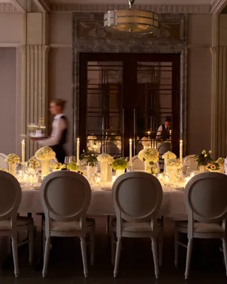 Elegant private dining room with a long candlelit table, floral centrepieces, and staff preparing place settings.