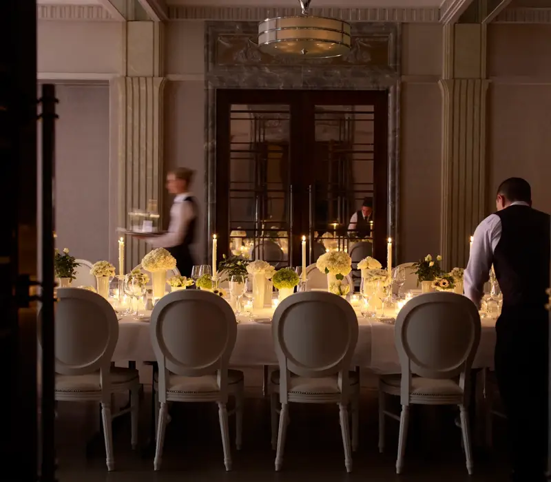 Elegant private dining room with a long candlelit table, floral centrepieces, and staff preparing place settings.