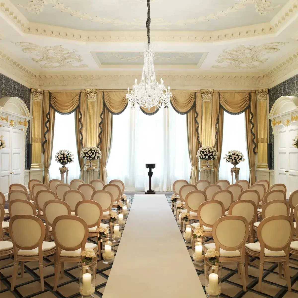 Elegant ballroom set for a wedding ceremony with cream chairs, floral arrangements, candlelit aisle, and a crystal chandelier