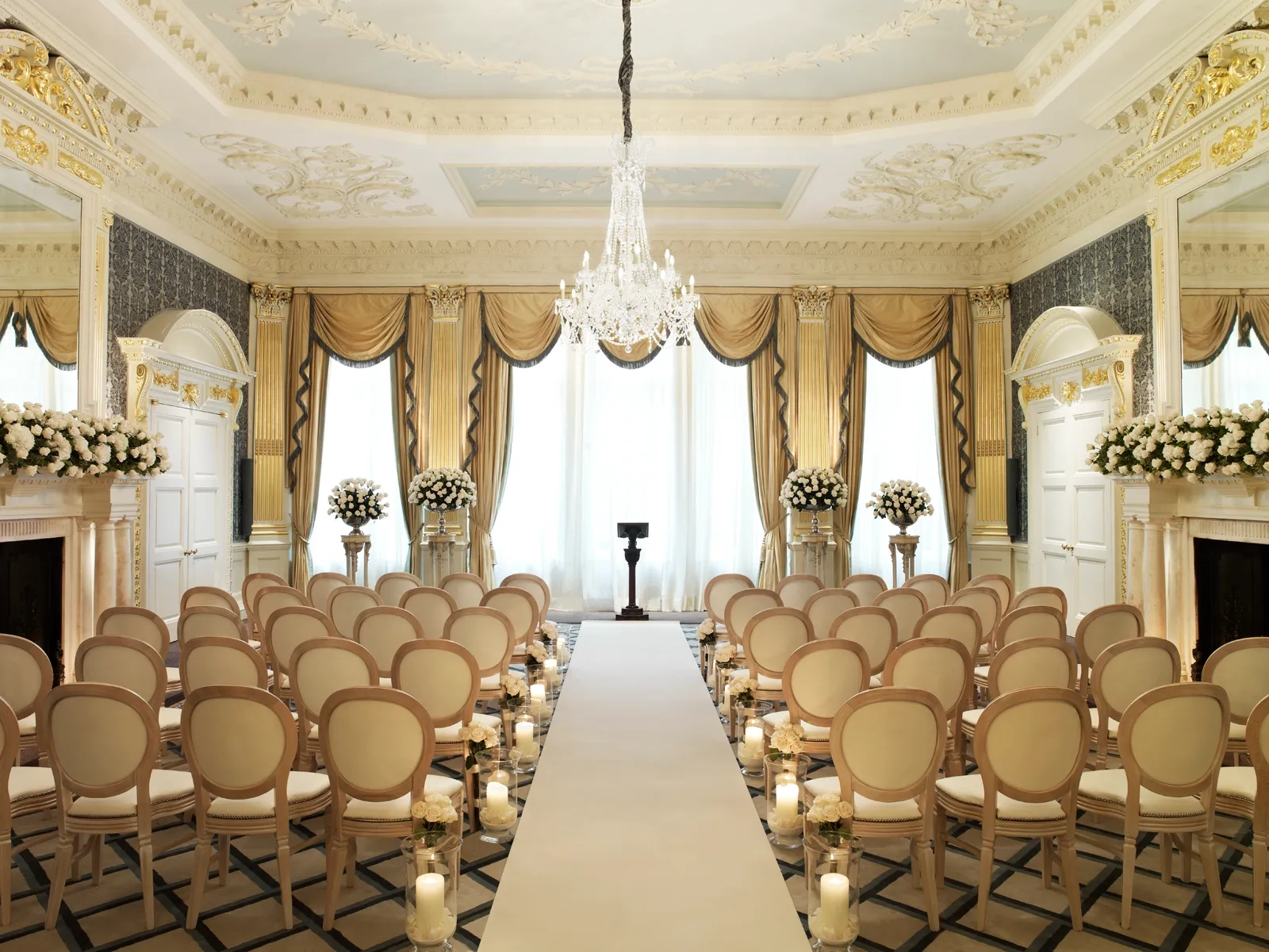 Elegant ballroom set for a wedding ceremony with cream chairs, floral arrangements, candlelit aisle, and a crystal chandelier