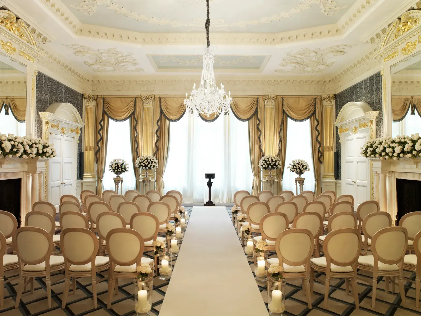 Elegant ballroom set for a wedding ceremony with cream chairs, floral arrangements, candlelit aisle, and a crystal chandelier