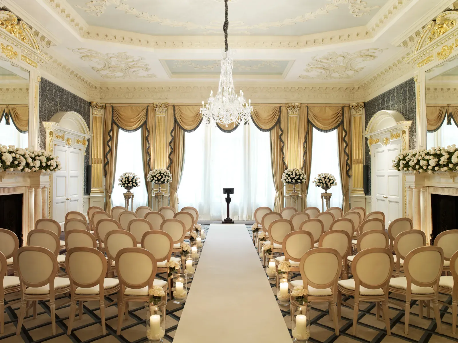 Elegant ballroom set for a wedding ceremony with cream chairs, floral arrangements, candlelit aisle, and a crystal chandelier