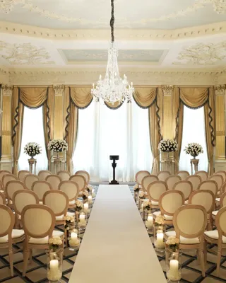 Elegant ballroom set for a wedding ceremony with cream chairs, floral arrangements, candlelit aisle, and a crystal chandelier