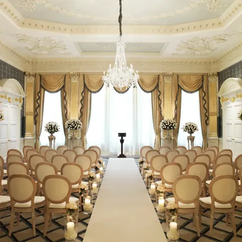 Elegant ballroom set for a wedding ceremony with cream chairs, floral arrangements, candlelit aisle, and a crystal chandelier