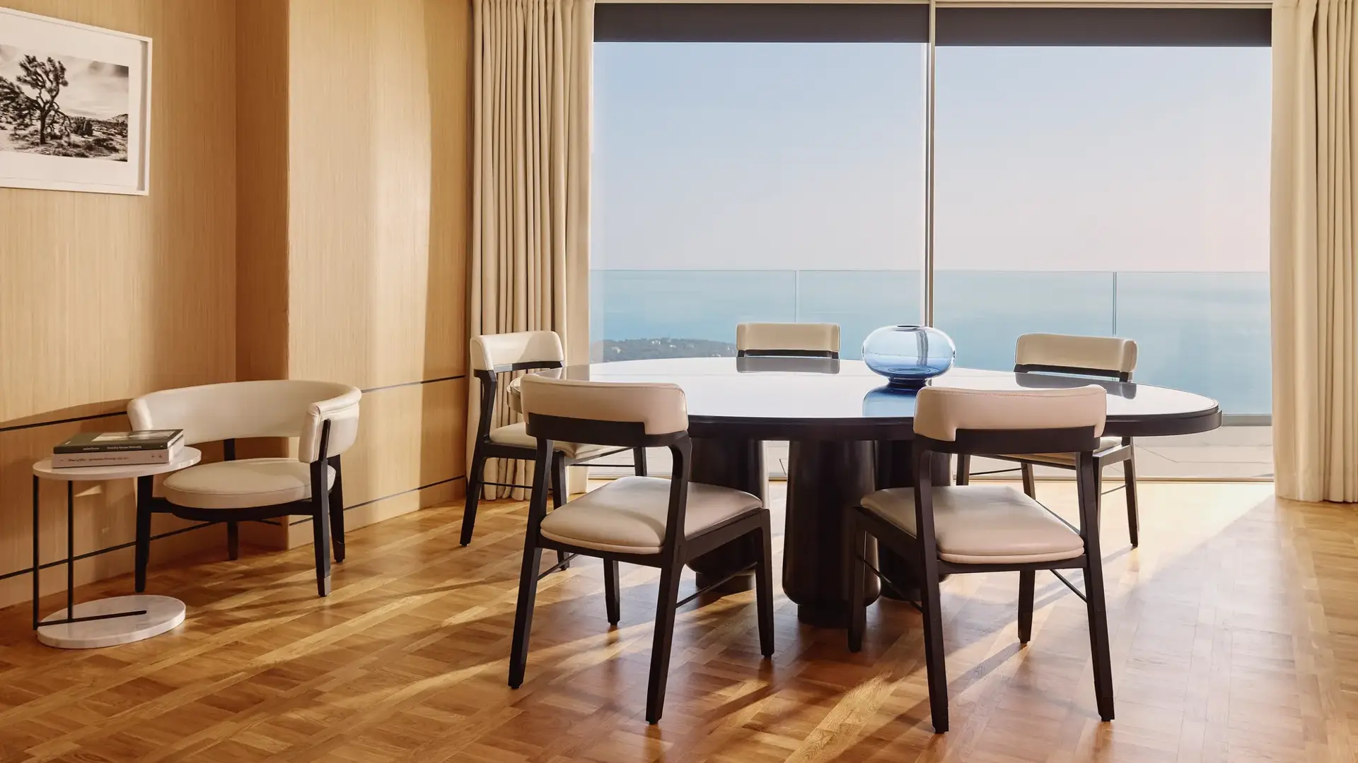 Elegant dining room with round table, modern chairs, and large windows offering sea views, accented by light wood panelling and soft natural light.