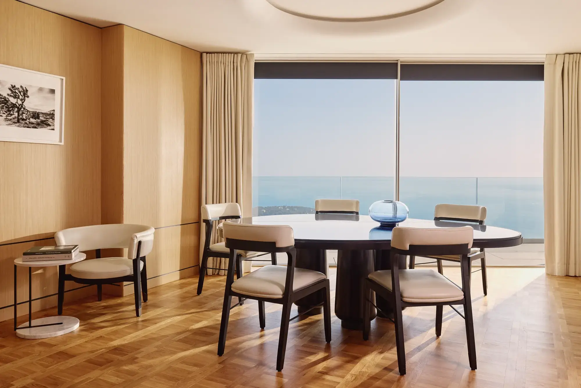 Elegant dining room with round table, modern chairs, and large windows offering sea views, accented by light wood panelling and soft natural light.