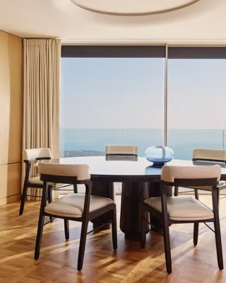 Elegant dining room with round table, modern chairs, and large windows offering sea views, accented by light wood panelling and soft natural light.