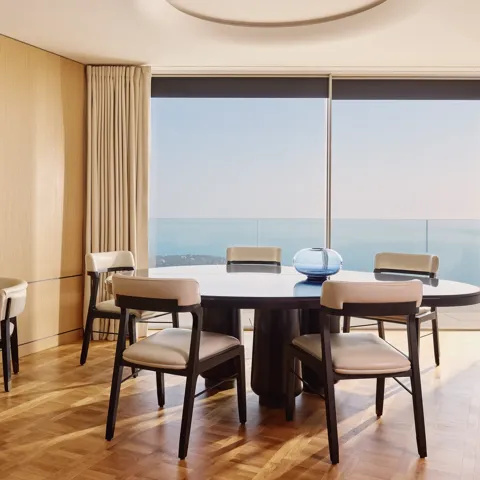 Salle à manger élégante avec table ronde, chaises modernes et grandes baies vitrées donnant sur la mer, rehaussée de panneaux en bois clair et de lumière naturelle.