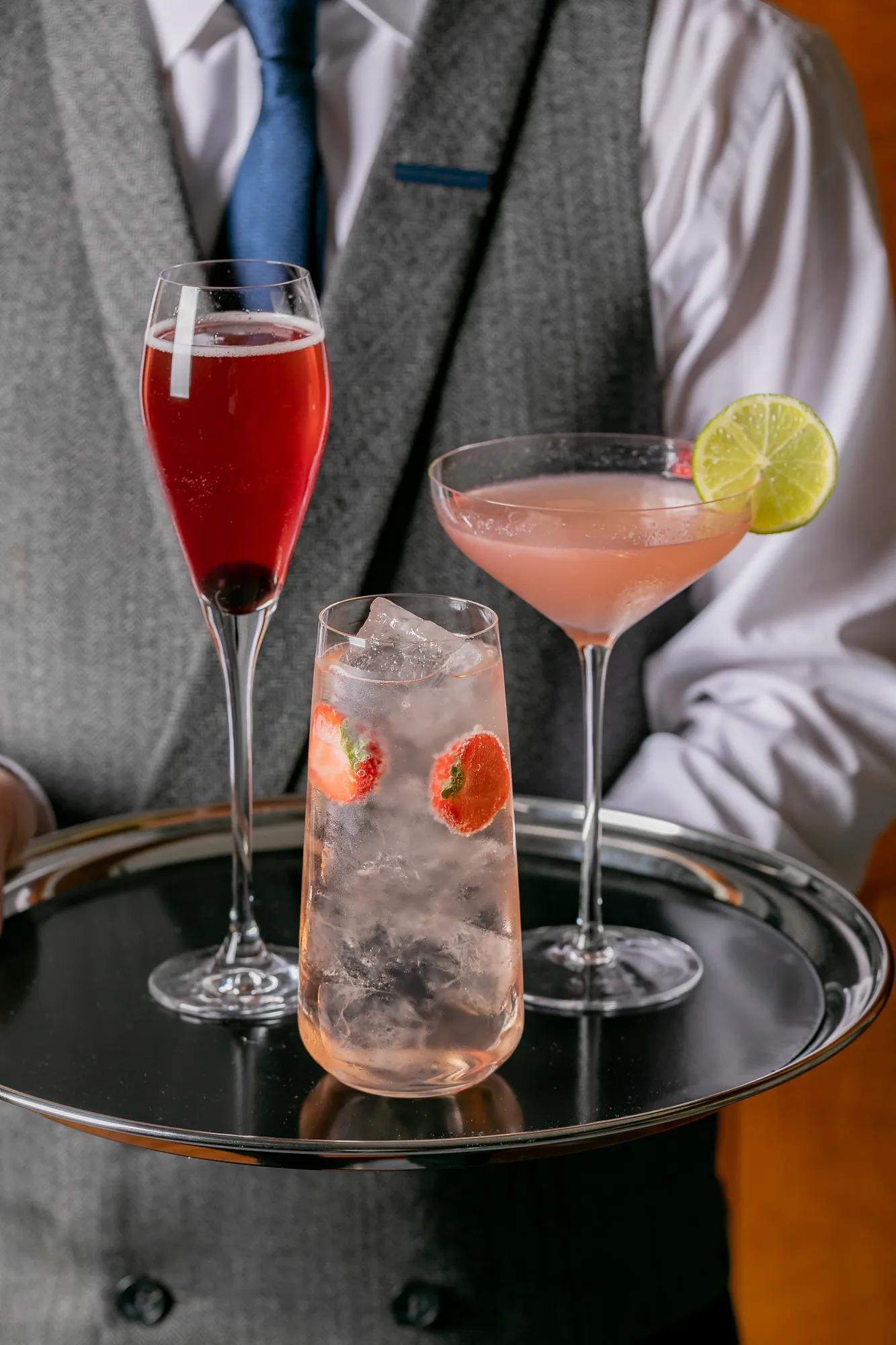 Waiter holding silver tray with three colorful cocktails, including strawberry spritz and martini with lime.