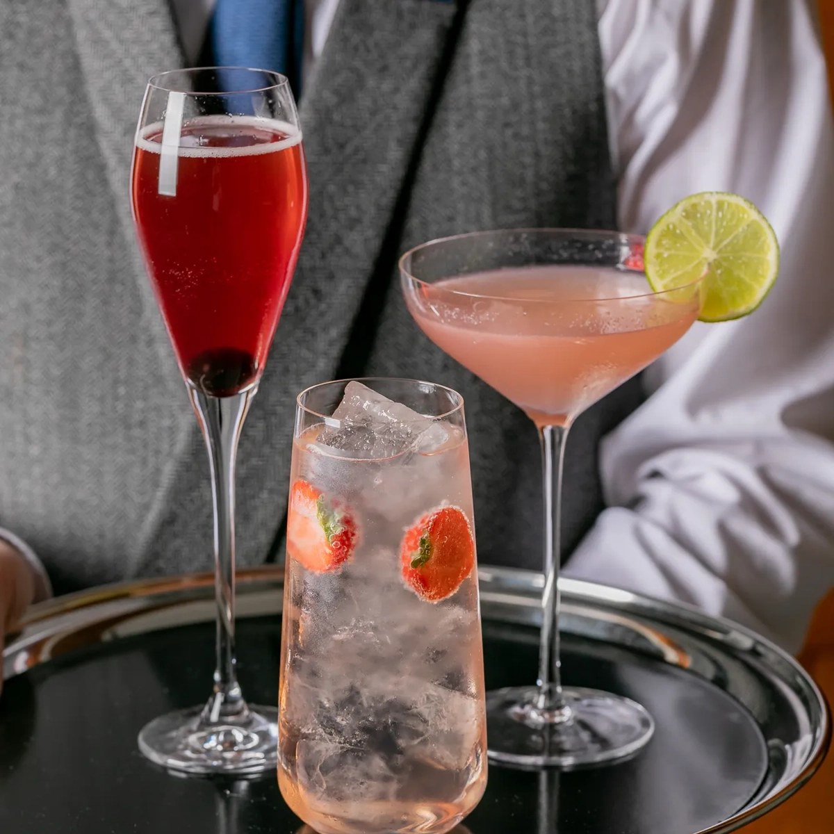 Waiter holding silver tray with three colorful cocktails, including strawberry spritz and martini with lime.