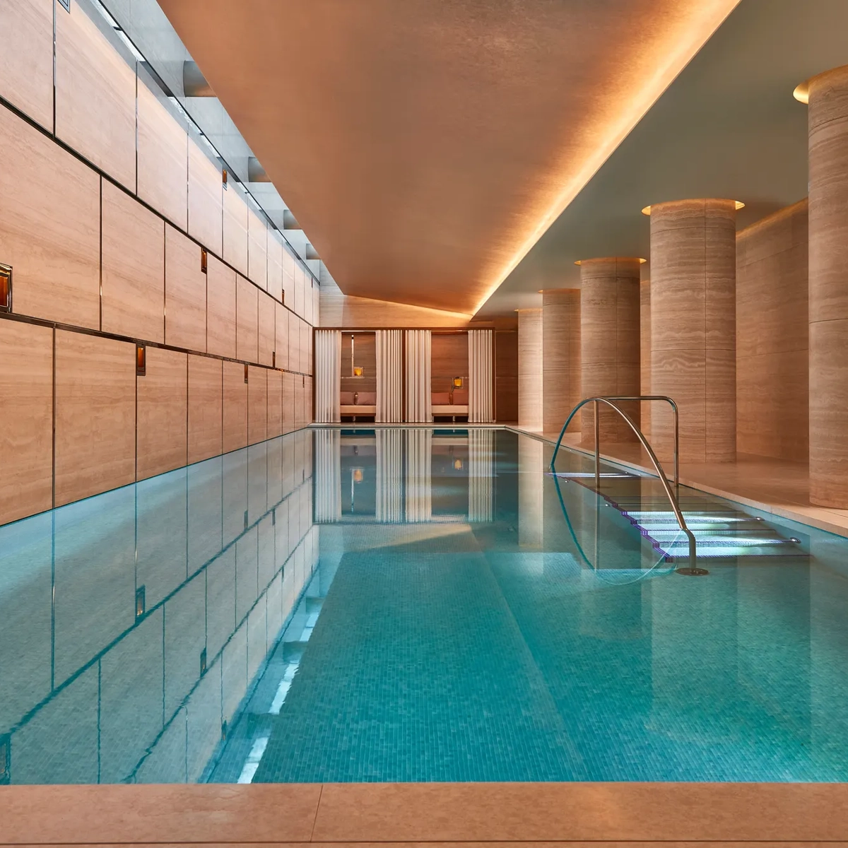 Indoor swimming pool with turquoise tiles, soft lighting, and stone columns, leading to curtained cabanas at the far end.