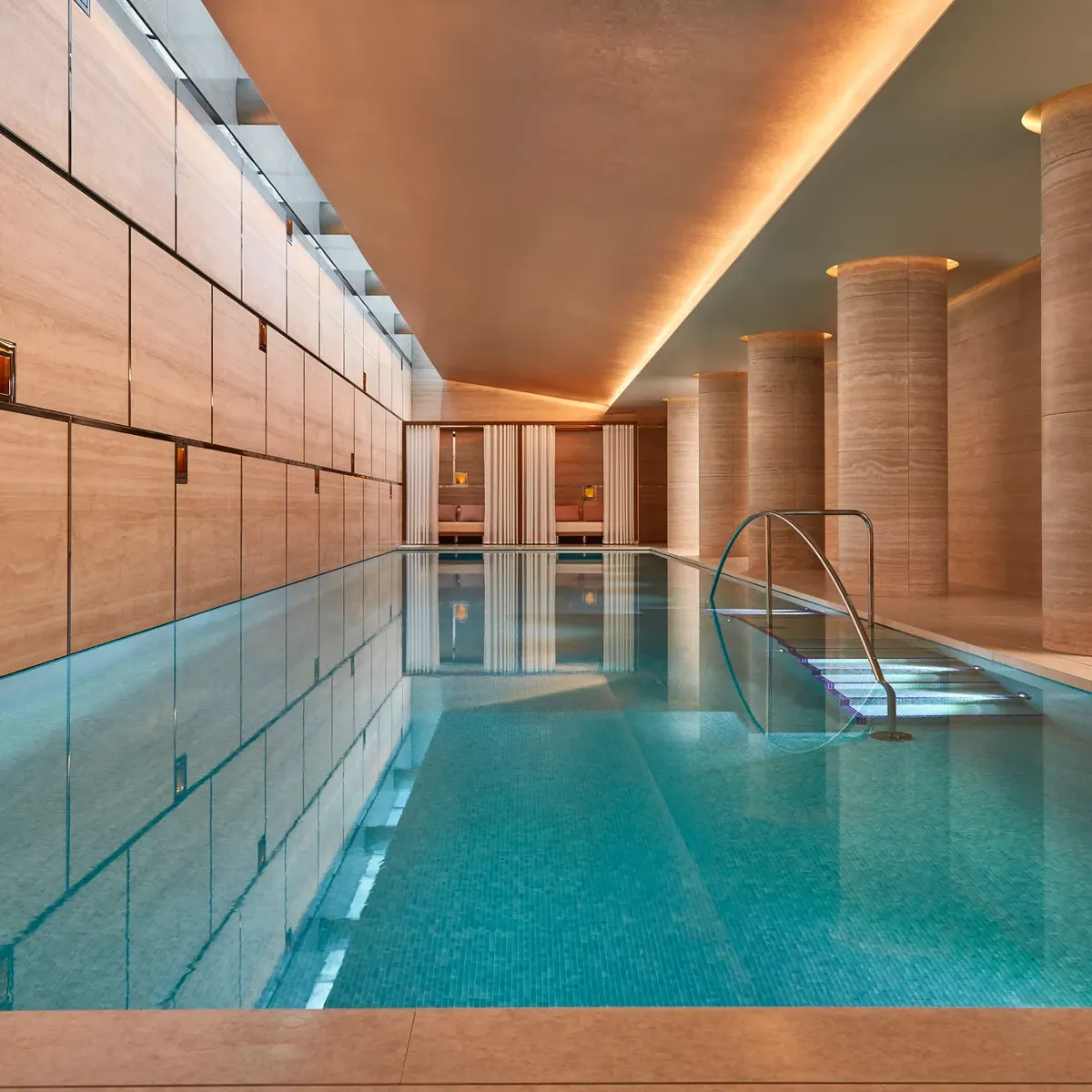 Indoor swimming pool with turquoise tiles, soft lighting, and stone columns, leading to curtained cabanas at the far end.