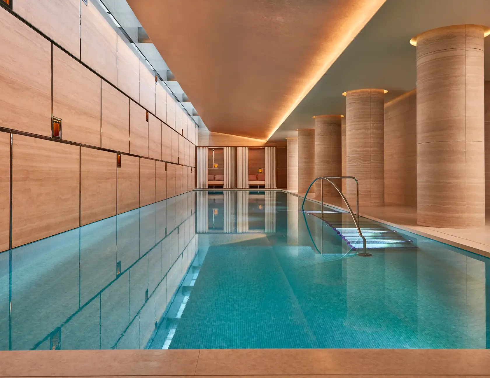 Indoor swimming pool with turquoise tiles, soft lighting, and stone columns, leading to curtained cabanas at the far end.