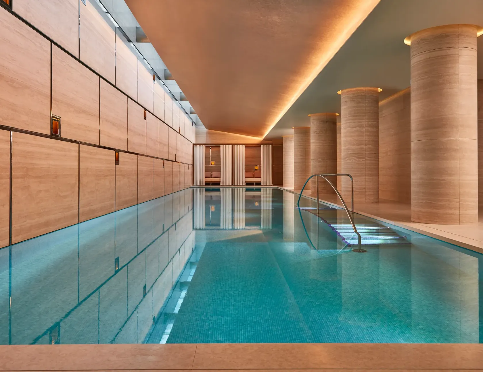 Indoor swimming pool with turquoise tiles, soft lighting, and stone columns, leading to curtained cabanas at the far end.