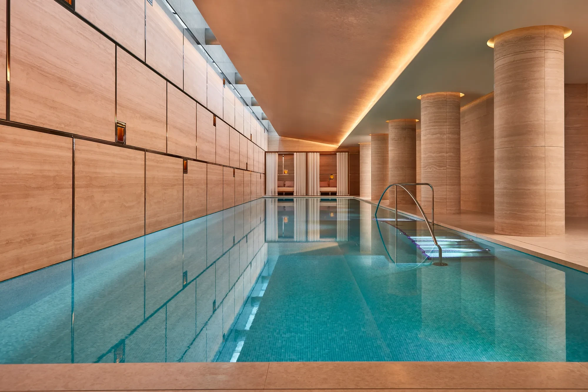 Indoor swimming pool with turquoise tiles, soft lighting, and stone columns, leading to curtained cabanas at the far end.