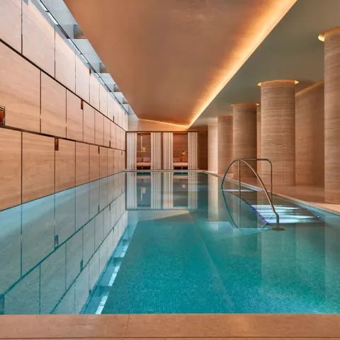 Indoor swimming pool with turquoise tiles, soft lighting, and stone columns, leading to curtained cabanas at the far end.