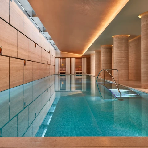 Indoor swimming pool with turquoise tiles, soft lighting, and stone columns, leading to curtained cabanas at the far end.
