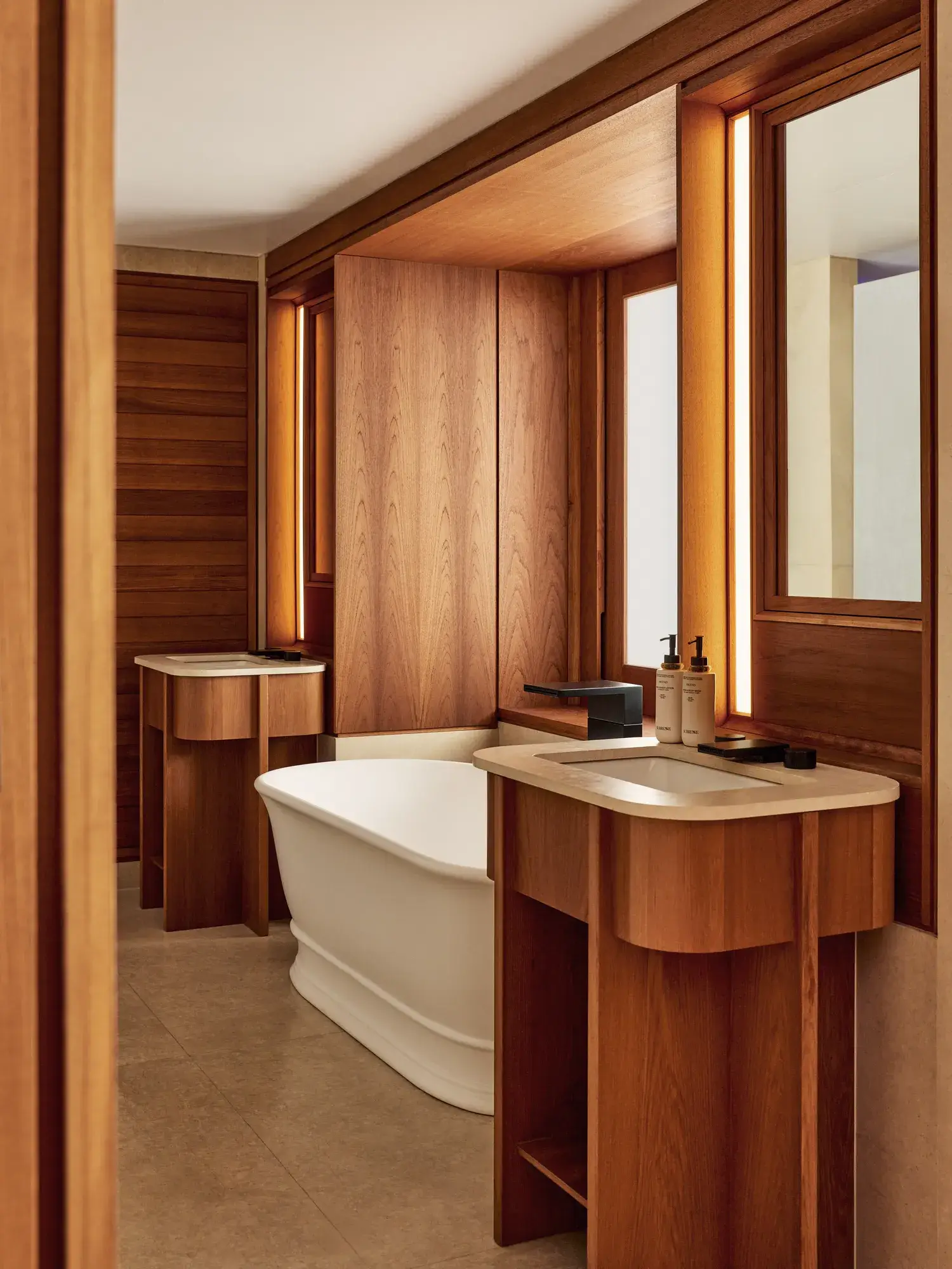 Luxurious bathroom in warm stone with wooden cabinetry, a freestanding bathtub, twin vanities, and backlit mirrors.