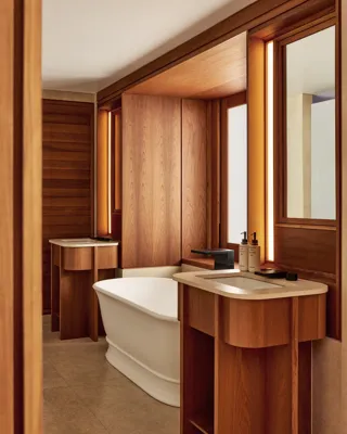 Luxurious bathroom in warm stone with wooden cabinetry, a freestanding bathtub, twin vanities, and backlit mirrors.