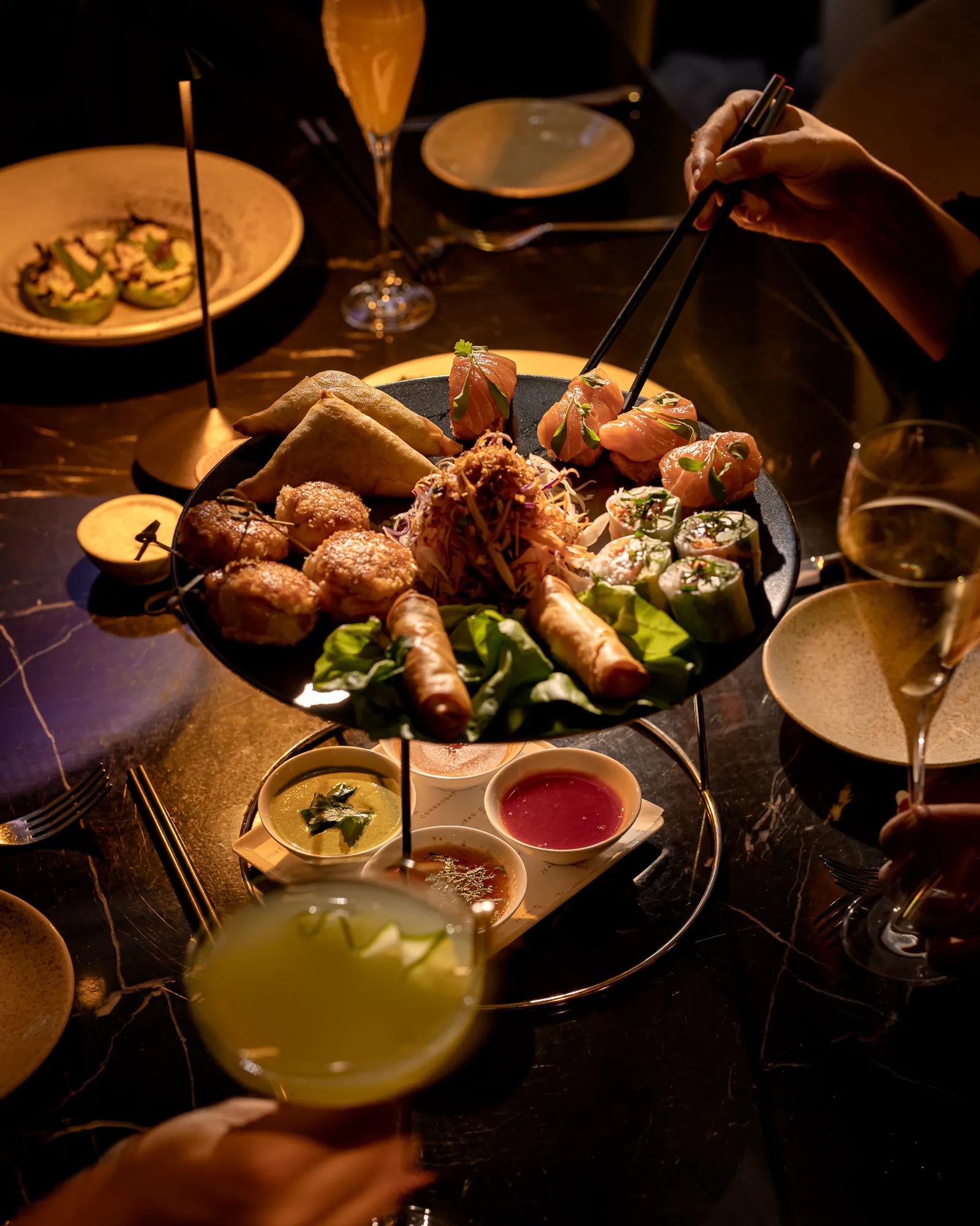 Sharing platter of sushi, spring rolls and appetisers served with dipping sauces and cocktails on a dining table.