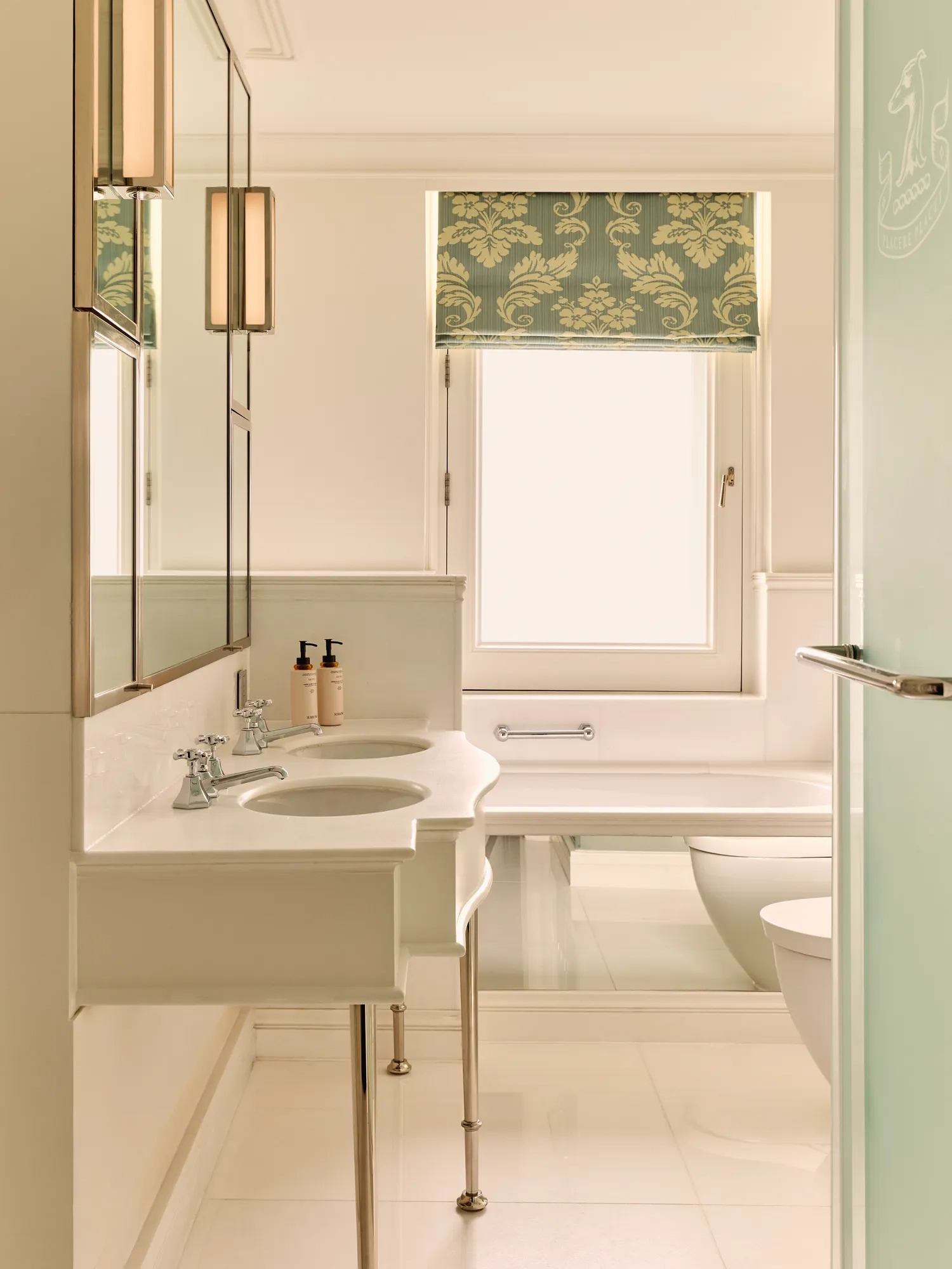 Light, elegant bathroom with twin sinks, a bathtub, and a patterned green window shade.