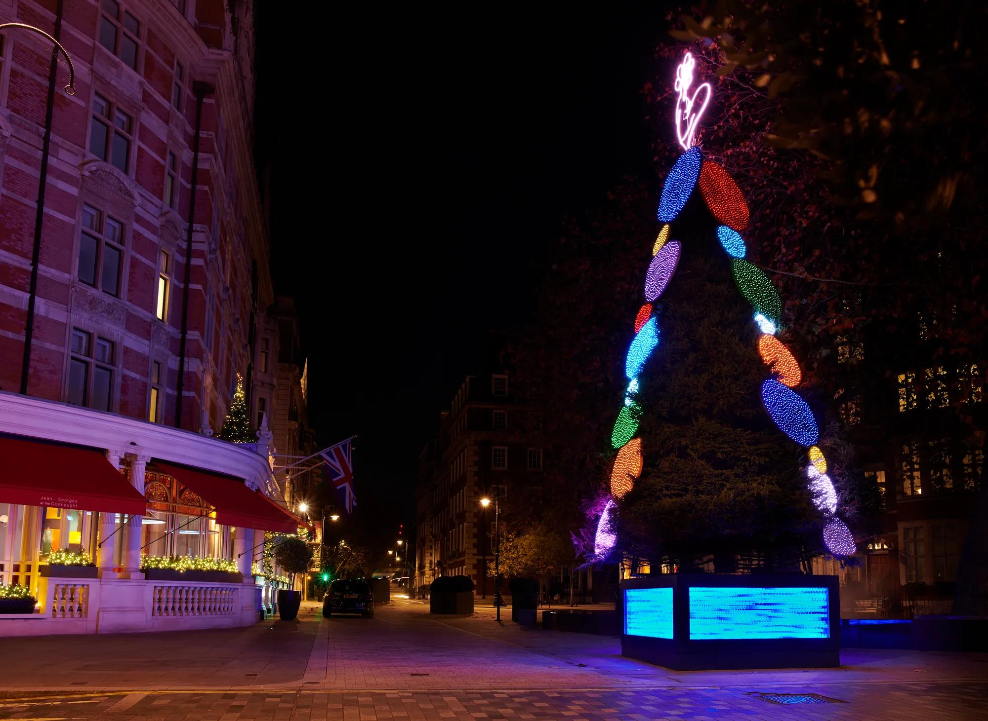 Christmas tree with colourful light ornaments and neon topper outside luxury hotel at night.