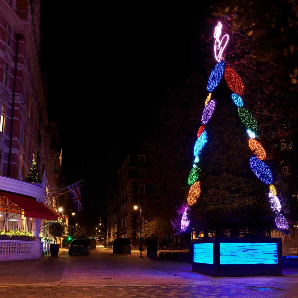 Christmas tree with colourful light ornaments and neon topper outside luxury hotel at night.
