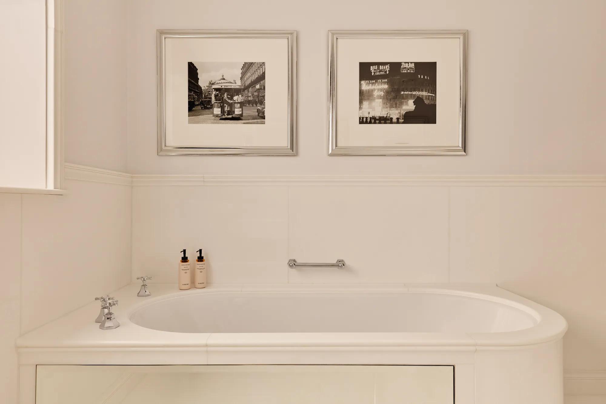 Cream bathroom with freestanding bathtub, chrome taps, toiletries, and two framed black-and-white photos above.