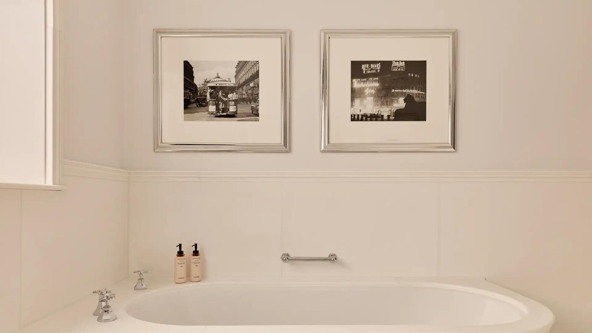 Cream bathroom with freestanding bathtub, chrome taps, toiletries, and two framed black-and-white photos above.