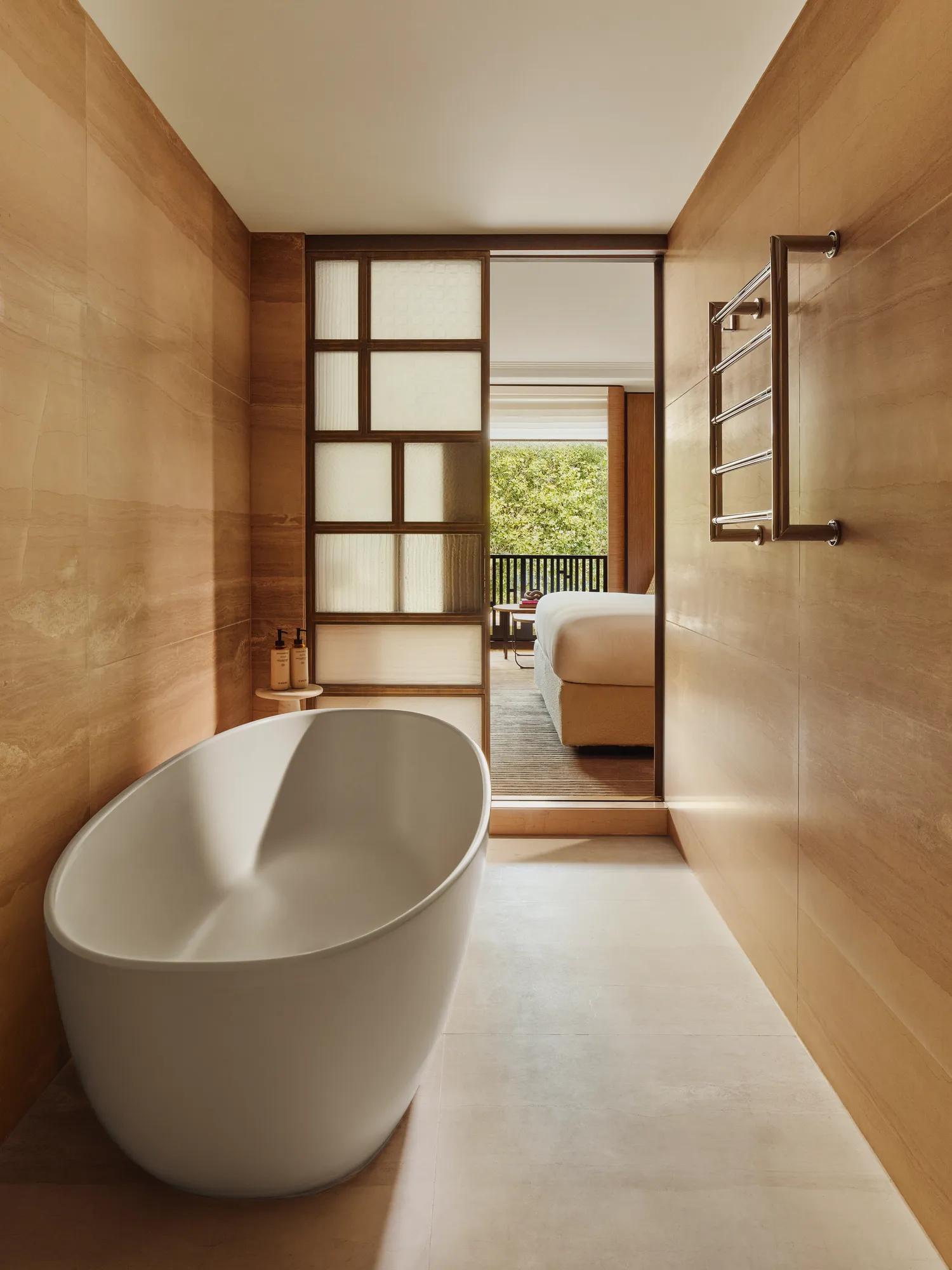Freestanding oval bathtub in a wood-panelled bathroom, with sliding screen opening to a bedroom and balcony views.