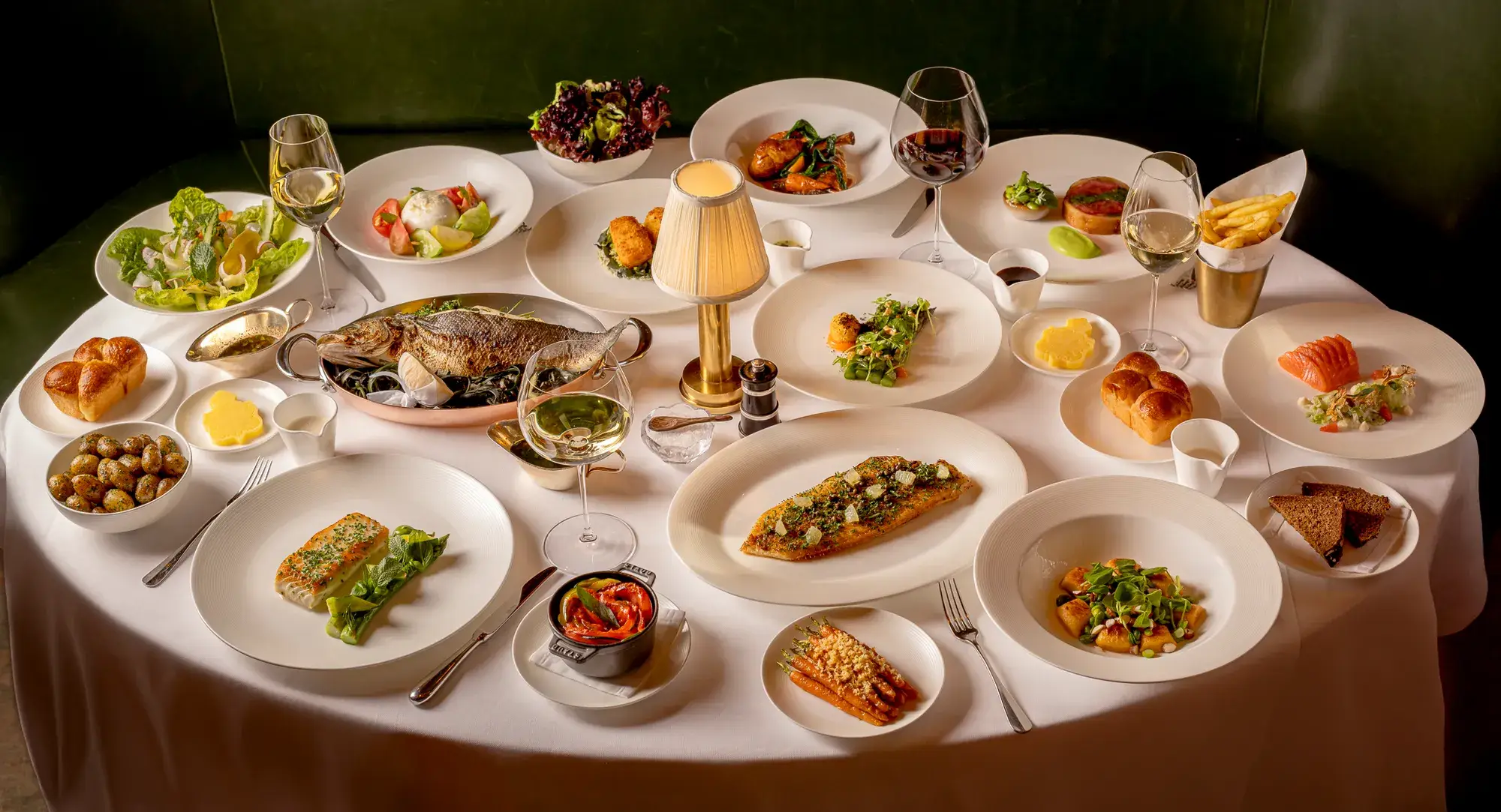 Table set with assorted gourmet dishes, salads, fish, sides, and wine glasses around a central lamp on a white tablecloth