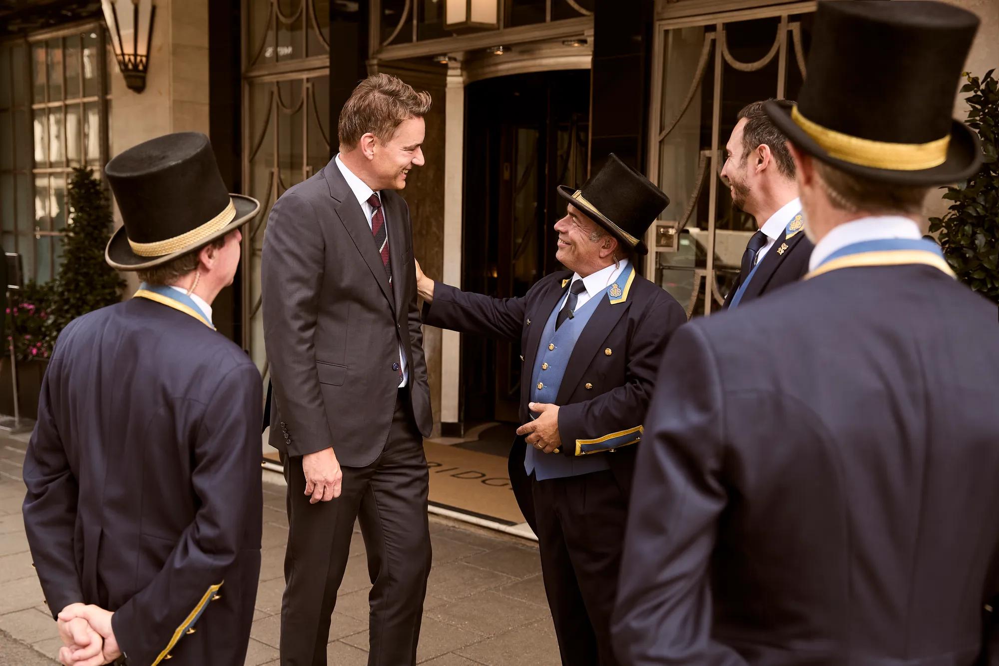 A hotel guest in a suit warmly greeted by Claridge’s doormen wearing navy uniforms with gold trim and top hats, standing outside the hotel entrance and sharing a friendly conversation.