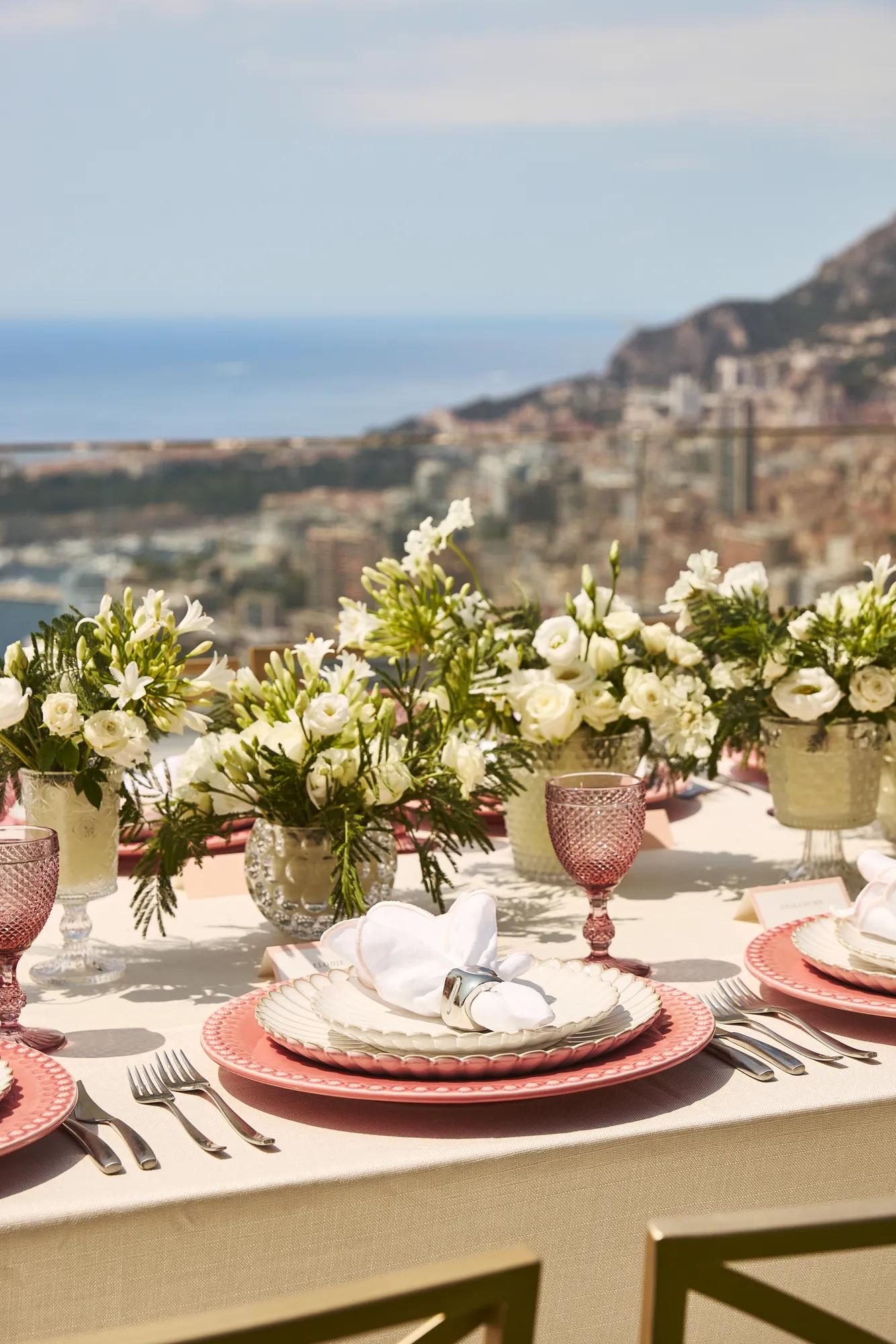 Gros plan sur une élégante table en extérieur avec des assiettes roses, des serviettes blanches, de la verrerie en cristal et des arrangements floraux blancs, avec vue sur Monaco et la mer.
