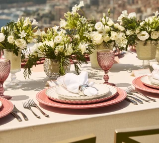 Gros plan sur une élégante table en extérieur avec des assiettes roses, des serviettes blanches, de la verrerie en cristal et des arrangements floraux blancs, avec vue sur Monaco et la mer.