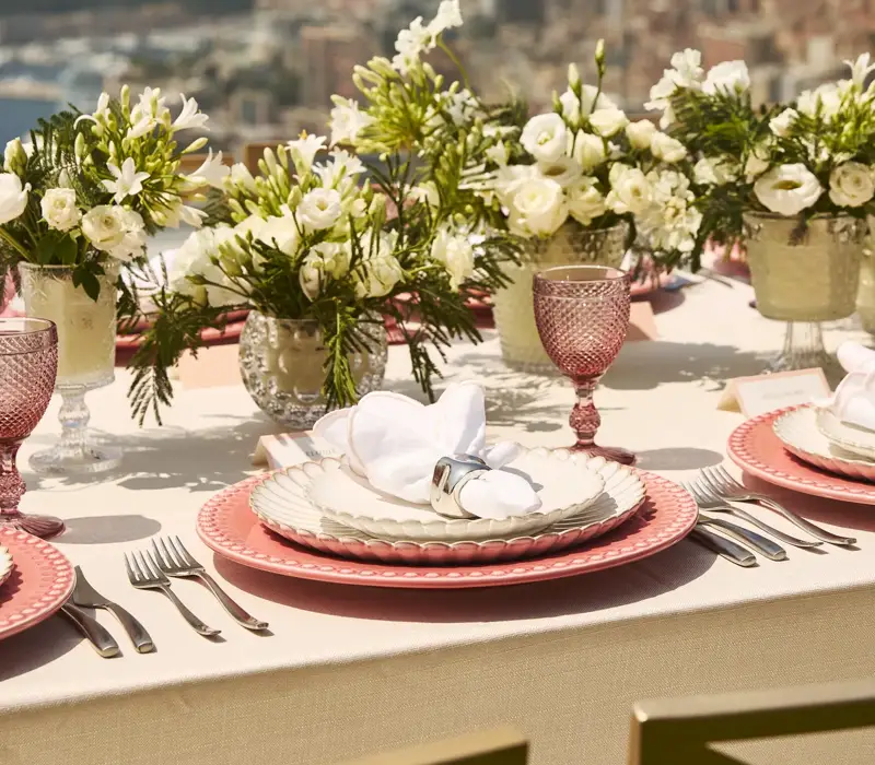 Gros plan sur une élégante table en extérieur avec des assiettes roses, des serviettes blanches, de la verrerie en cristal et des arrangements floraux blancs, avec vue sur Monaco et la mer.
