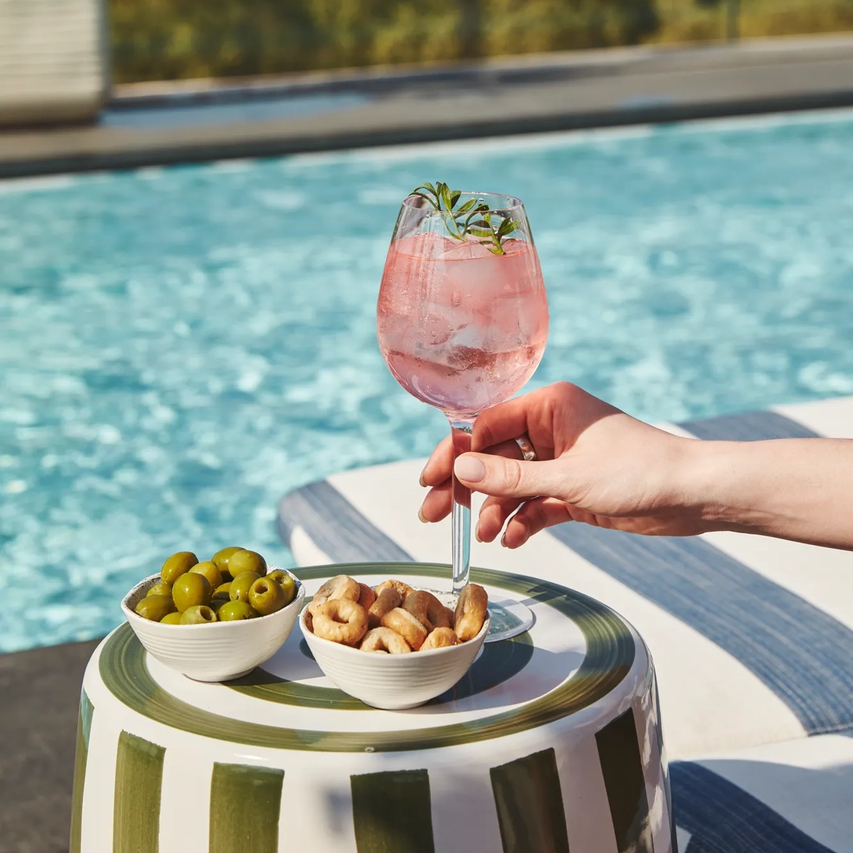 Hand holding pink cocktail with rosemary by poolside, beside bowls of olives and snacks on striped table