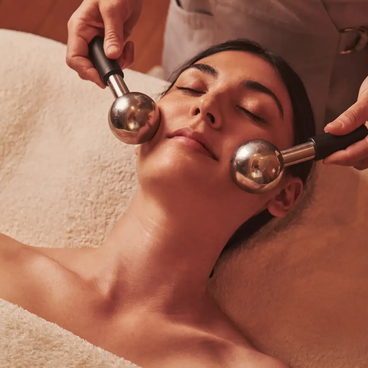 Person receiving a facial massage with two metal massage globes while lying wrapped in a soft towel.