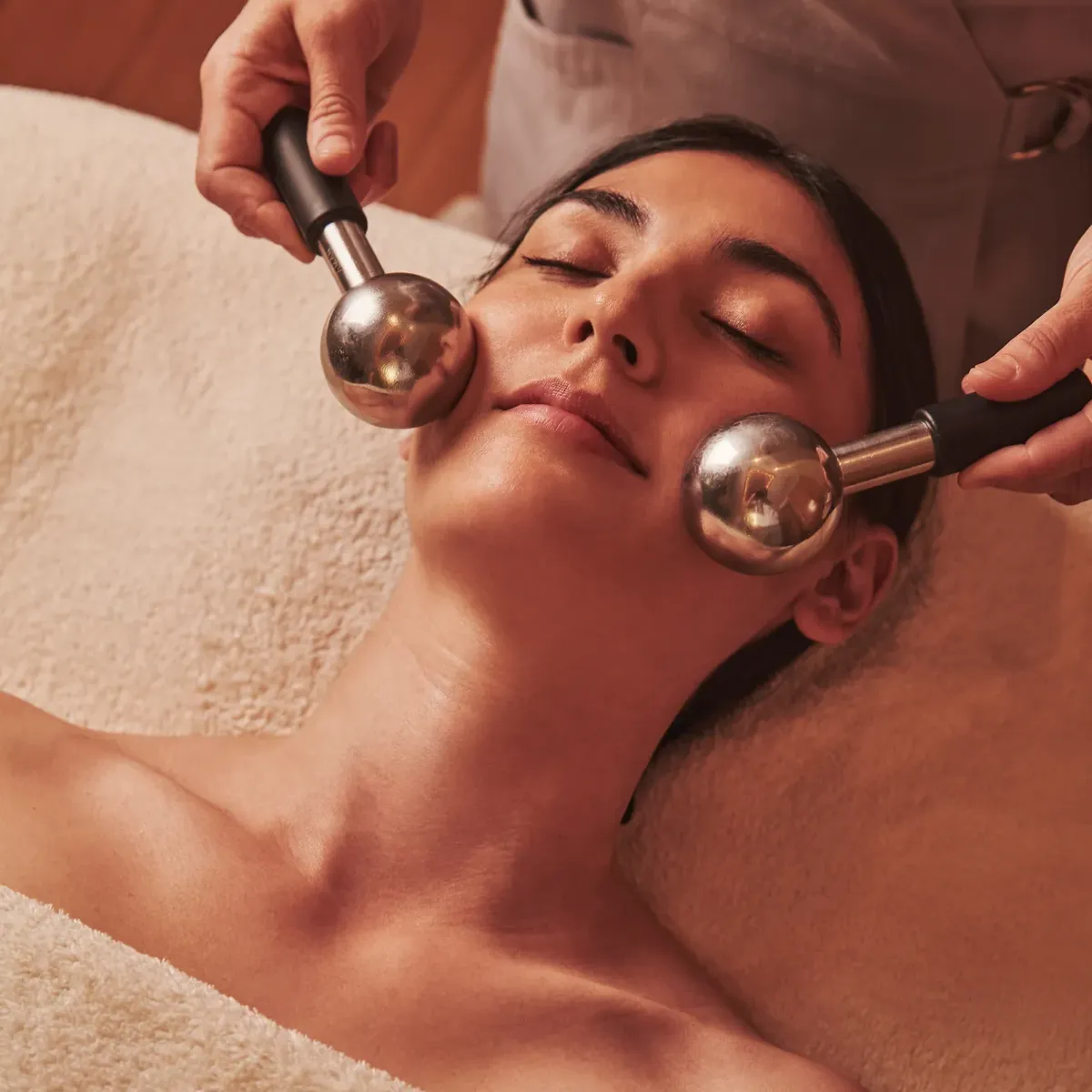 Person receiving a facial massage with two metal massage globes while lying wrapped in a soft towel.